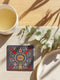 A square ceramic drink coaster with a smooth, matte charcoal grey finish. The surface features a symmetrical, intricate design in metallic gold, deep garnet red, and light grey. In the center is a golden All-Seeing Eye with a red iris, radiating light. The eye is framed by a golden circle and flanked by botanical elements, including red pomegranates and grey flowers with gold details. The design has a folk art, gothic botanical style.